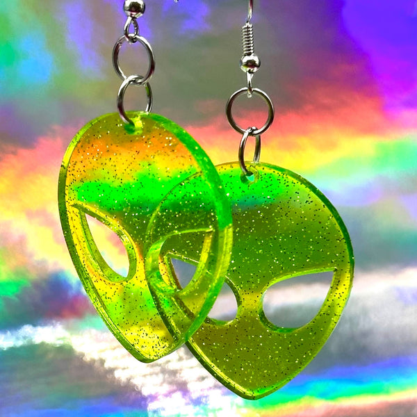 Neon green transparent acrylic with silver glitter alien head-shaped dangle earrings on a rainbow background