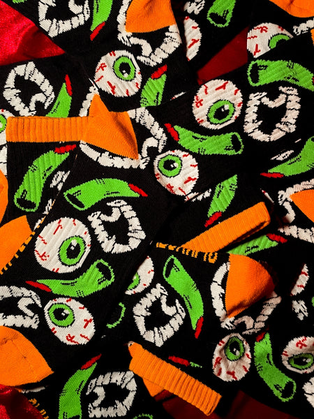 black cotton crew socks with orange cuffs, heels, and toes with an all-over pattern of plastic vampire teeth, witch fingers, and green bloodshot eyeballs. Shown flat in pile of socks