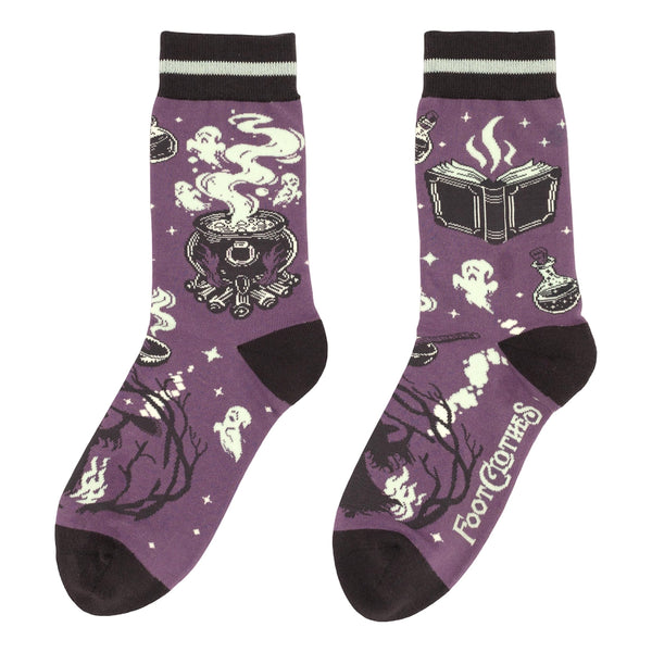 unisex cotton blend crew socks in medium purple with black toes, heels, and black & cream striped cuffs with purple, black, and cream design of spell books, bubbling cauldrons, bottles of potions, ghosts, and a witch holding a broom. Shown flat from opposite side