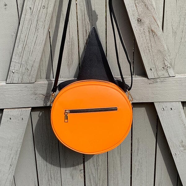 Faux leather shoulder bag in the shape of an orange jack-o’-lantern pumpkin with smiling face and black witch hat with yellow stars. Bag has adjustable black faux leather strap. Shown from front hanging on fence from back to display solid orange back with zippered compartment