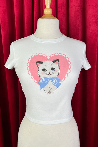 White cropped t-shirt with a graphic of a black and white kitten with a light blue bow around its neck in front of a pink scalloped heart on a mannequin against a red curtain background