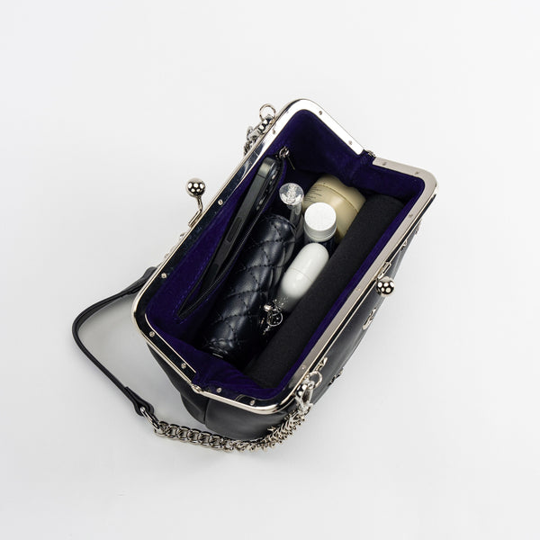 black textured faux leather purse with silver metal kiss lock and silver metal stars, crescent moons, and sun embellishments. Attached is an 11" chain link and faux leather shoulder strap. Shown open from top to display purple-blue velvet lining