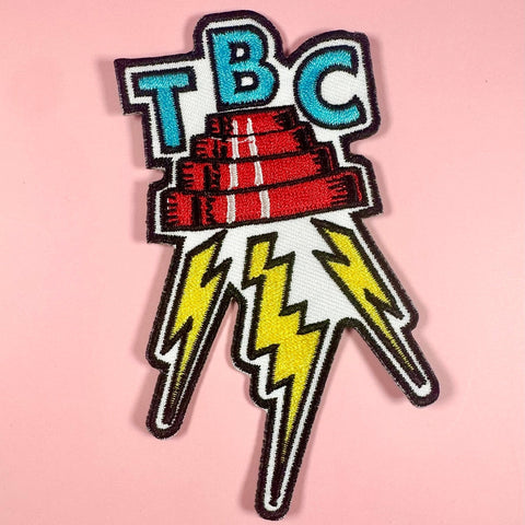 embroidered patch with 'TBC' lettering in blue and lightning bolts in between a DEVO energy dome on a pink background