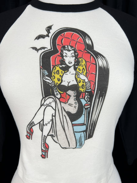 off-white crop length black raglan shoulder top with round neck, three quarter sleeves, and printed illustration of pinup style woman with stitched legs wearing gown and sitting in coffin-style large chair. Printed in red, black, blue, and yellow. Shown on dress form in close up of illustration