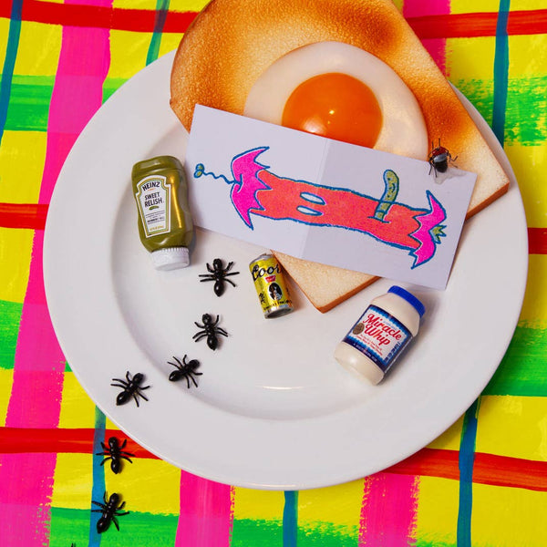 heavyweight card stock note card with a neon pink, coral, green, and blue risograph image of a rotten apple core with worm that wraps around both exterior sides of card. Shown flat on plate alongside plastic ants, fly, and egg with toast on neon plaid background