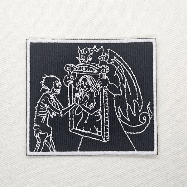 embroidered black twill patch with white border featuring white woodcut-style art of a devil holding a mirror to a skeleton and showing them the reflection of a woman. Shown flat