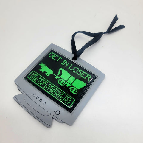 Holiday ornament made of matte grey laser cut acrylic in the shape of a PC computer monitor with domed resin screen displaying art from the Oregon Trail video game with "Get in loser, we're going to die of dysentery" message in neon green UV reactive paint. Ornament has black satin ribbon hanger. Shown flat from front