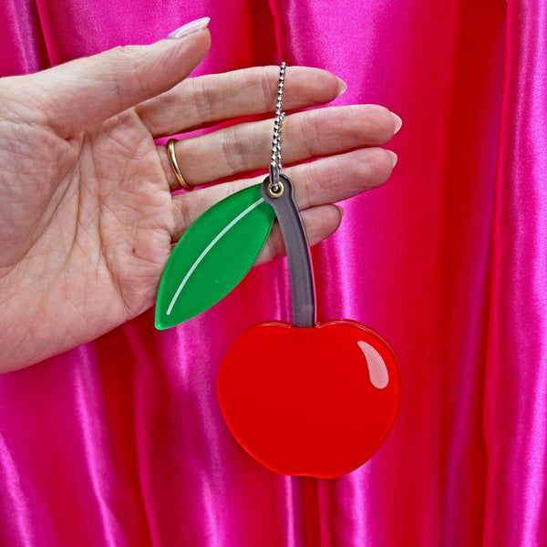 red acrylic cherry mirror with brown stem and green leaf on ball chain shown slid closed held in front of a pink background