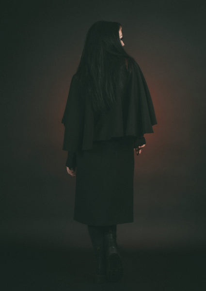 Model wearing black full length faux wool-blend coat with shawl collar and attached elbow-length cape. Showing back view