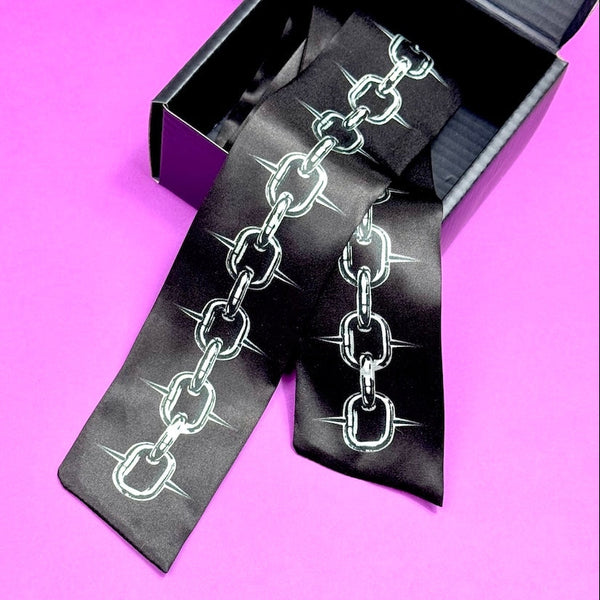 skinny black silk scarf with printed silver pattern of spiky link style chain shown in black packaging box on purple background