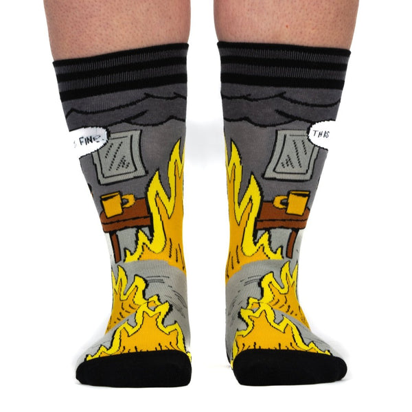 black, grey, yellow, and orange unisex crew socks with black and grey stripe cuffs and black toes/heels with all over pattern of a cartoon dog wearing a hat sitting at a kitchen table surrounded by flames with a speech bubble saying "THIS IS FINE." Shown worn from the front