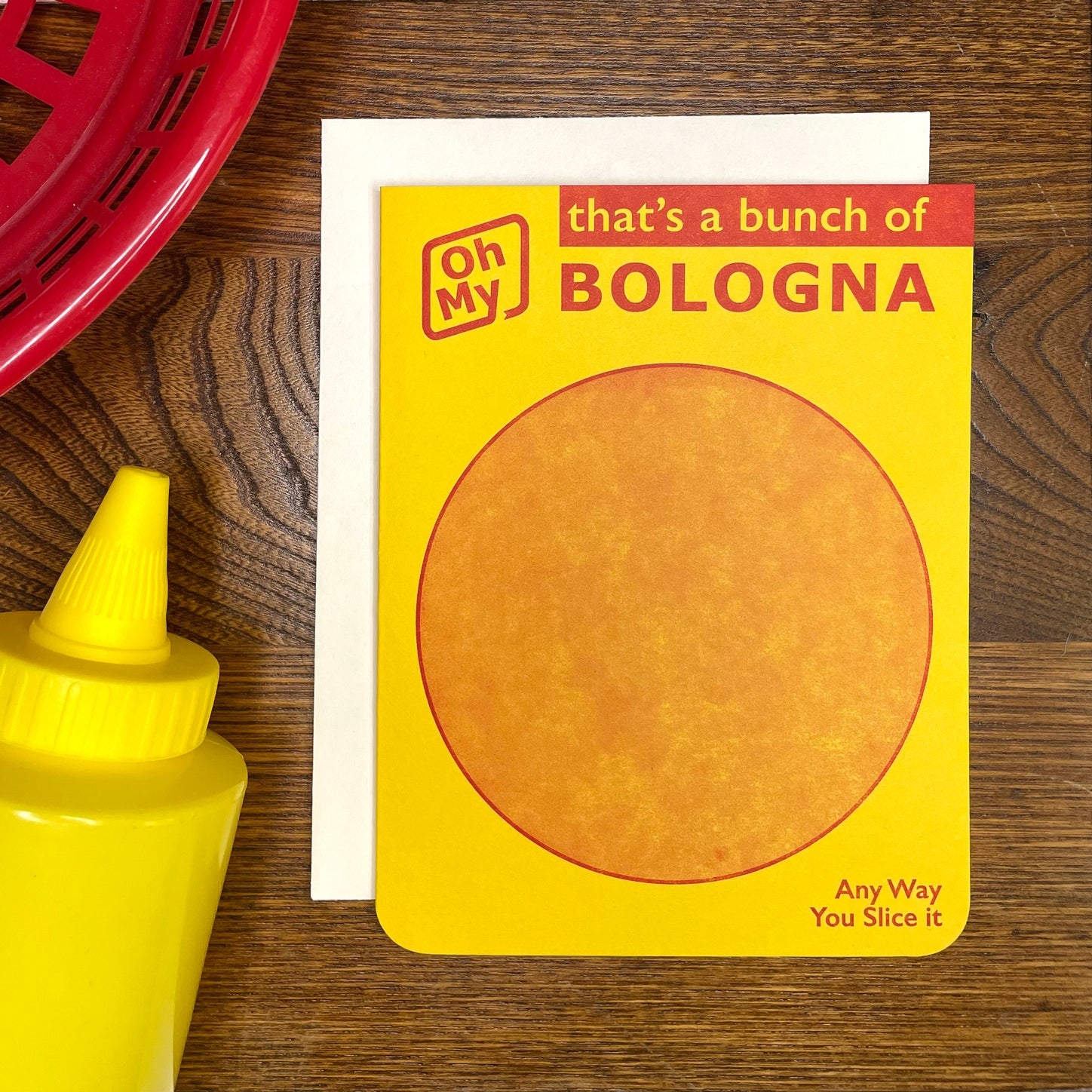 Bright yellow notecard resembling Oscar Meyer bologna package with round image of sandwich meat and message “Oh My” “that’s a bunch of BOLOGNA” “Any Way You Slice It”. Shown on wooden background with mustard squeeze bottle