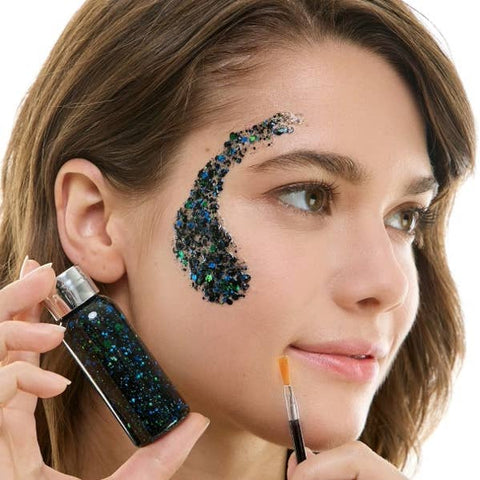 Woman applying black, green, and blue decorative glitter makeup with a brush on a white background