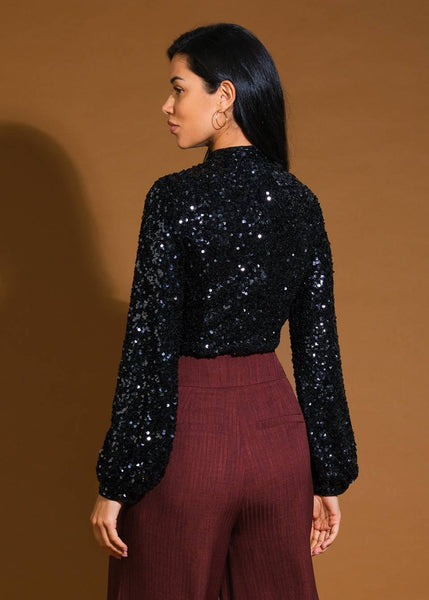 model wearing a black top with high neckline and long balloon-style sleeves in an allover black sequin fabric. Shown from  back
