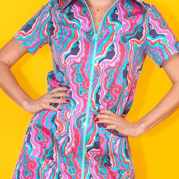 model wearing a cotton short sleeve shift mini dress with patch pockets and light blue plastic front zipper. Pattern of dress is faces in silhouette, lips, eyes, tiny platypuses, stars, eggs, and paisley in various shades of blue, aqua, red, and pink. Shown from front in close up