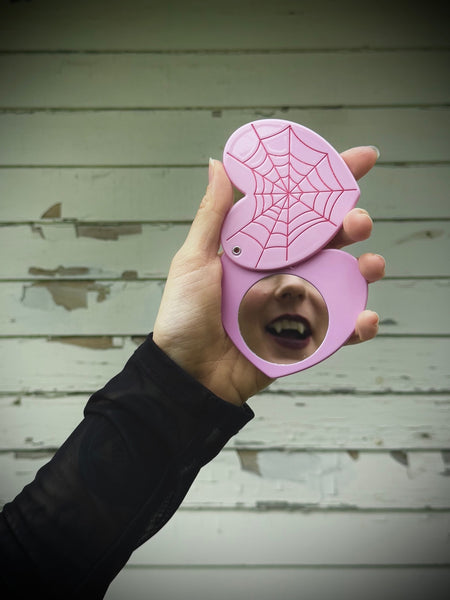 Hand holding a pink heart-shaped hand mirror with red spider web design against a wooden background and mouth with fangs shown in mirror