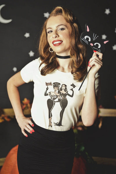 model wearing ivory white t-shirt with printed illustration of two pinup-style women holding masquerade masks, one dressed as the Bride of Frankenstein and the other dressed as a black cat. Shown from front