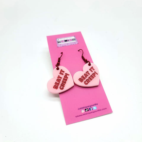 pink laser cut acrylic heart shaped dangle earrings resembling conversation heart candies with the message "BEAT IT CREEP!" in red capital letters. Shown on packaging card