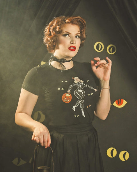 Model wearing a black short sleeve t shirt with a printed illustration of a pinup style woman wearing a skeleton costume holding a pumpkin basket surrounded by pairs of monster eyes
