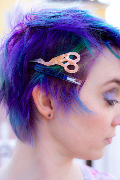 set of barrettes in the shape of stork scissors and seam ripper made of cellulose acetate shown in purple and green dyed short hair