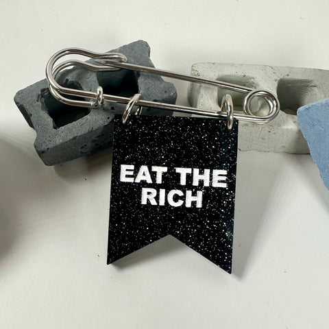 2" silver metal safety pin with acrylic black glitter banner attached with message "EAT THE RICH" in white