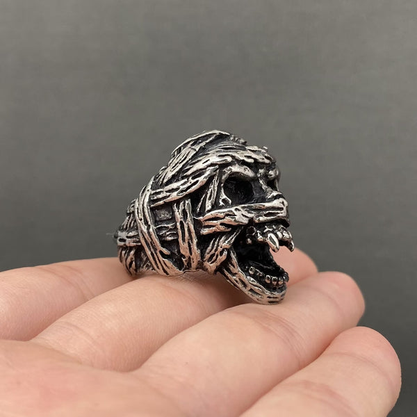 A silver stainless steel ring shaped like a mummy skull with snarling mouth, textured bandages and dark recessed details, shown resting on a person's fingers against a dark gray background. Shown from side
