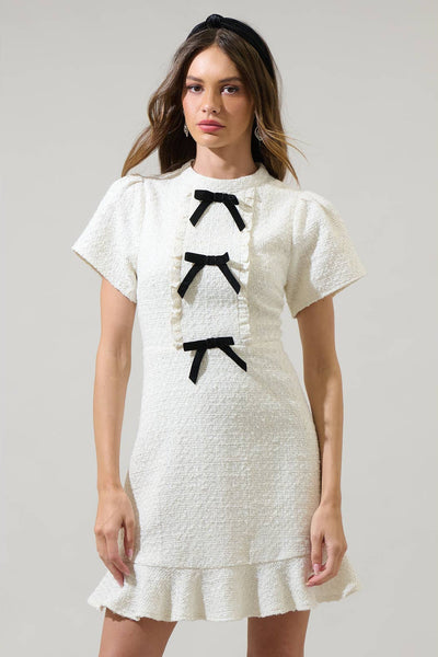model wearing white textured tweed woven mini dress with mock neck, short sleeves with slightly puffed shoulders, bodice with vertical ruffled bib style and three black bows running down the middle. Dress has a-line skirt ending above the knee in a wide gathered ruffle. Shown from the front