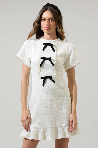 model wearing white textured tweed woven mini dress with mock neck, short sleeves with slightly puffed shoulders, bodice with vertical ruffled bib style and three black bows running down the middle. Dress has a-line skirt ending above the knee in a wide gathered ruffle. Shown from the front