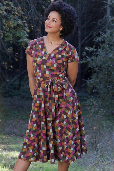 A woman wearing a stretchy cotton knit cap-sleeve dress with a black background rectangular grid in shades of pink, green, orange, and robin’s egg blue, with faux wrap surplice bodice and self tie sash. Shown standing outdoors on a gravel path with trees in the background.