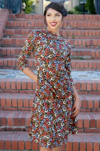 Model wearing 3/4 sleeve dress with slash neckline and all-over pattern of red, cream, and periwinkle violets on a black background. It has a slightly flared below the knee skirt and ruching on the left hip. Shown from the front 