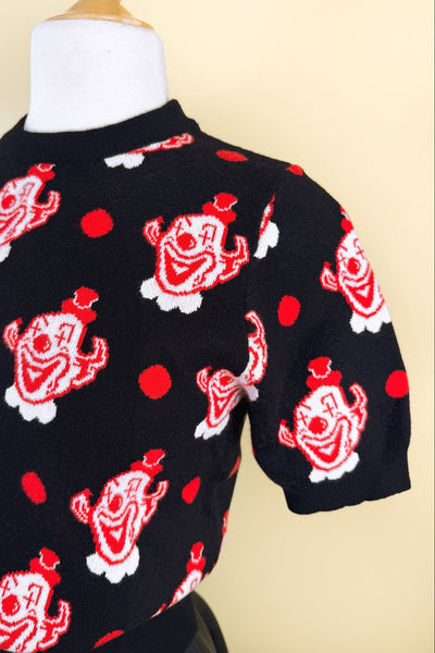 short sleeve acrylic knit sweater with black background and all-over tessellated white and red clown head print alternated with red polka dots. The top has a crew neck and ribbed hem. Shown from front in close up of pattern