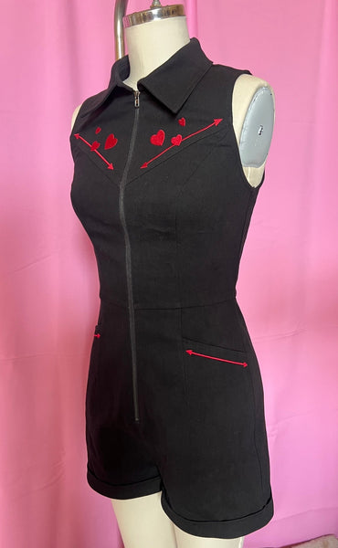 Black cotton blend stretch denim sleeveless romper with pointed collar, silver zipper running down the length of the piece, vertical seaming, red embroidered diagonal heart and arrow detail at the front yoke, red embroidered arrow detail below the diagonal pockets, and cuffed slightly flared shorts. Shown from three quarter angle on dress form