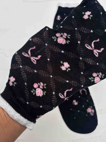 A pair of black crew socks with white trimmed cuffs and a pink rose and bow pattern on an openwork diamond pattern background shown in close up