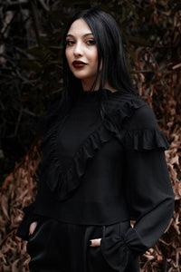 Model wearing a long sleeved black sheer jacquard chiffon blouse with a banded ruffled collar, deep ruffled yoke down the bodice, ruffles at the top of each full sleeve, and elasticized cuffs. Shown from a three quarter angle