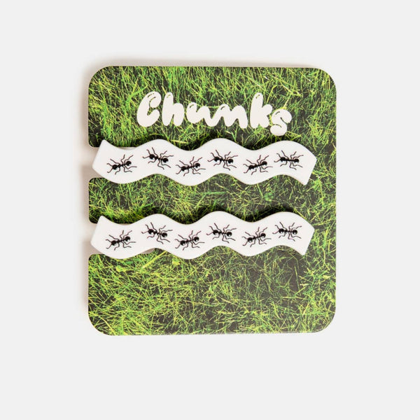 set of two bright white acetate alligator style hair clips in a wavy rectangle shape with painted detail of rows of ants along each clip. Shown on grass pattern backing card