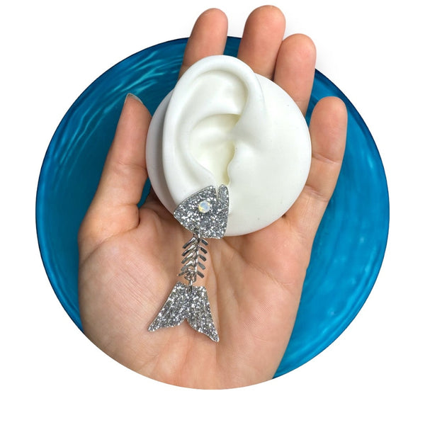 laser-cut silver glitter acrylic fish bone drop earrings with acrylic heads and tails with frosted rhinestone for each eye connected with bone-like chain. Single earring shown on mannequin ear