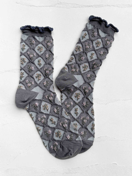 A pair of gray patterned socks with a ruffled elastic top, laid flat on a light textured background. The socks feature a diamond lattice pattern with small floral motifs in muted pink and green, and solid gray heels, toes, and cuffs. Shown flat on white background