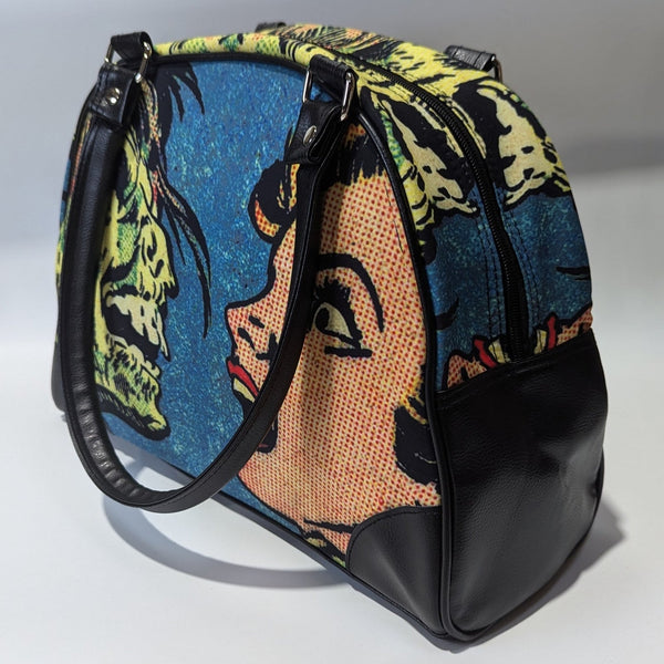 A bowler style purse with a satin body and printed image of comic book Ben Day style  art of a zombie face to face with a screaming woman. The purse has black faux leather handles, protective corners, sides, and bottom. Shown from side with handle hanging over side