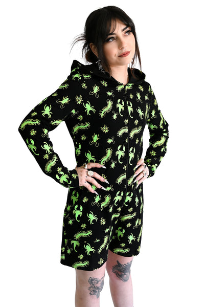 model wearing a black long sleeve hooded onesie with neon green insect pattern, front zip closure, and mid-length shorts. Shown from three quarter angle with hood down