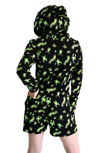 model wearing a black long sleeve hooded onesie with neon green insect pattern, front zip closure, and mid-length shorts. Shown from back with hood up