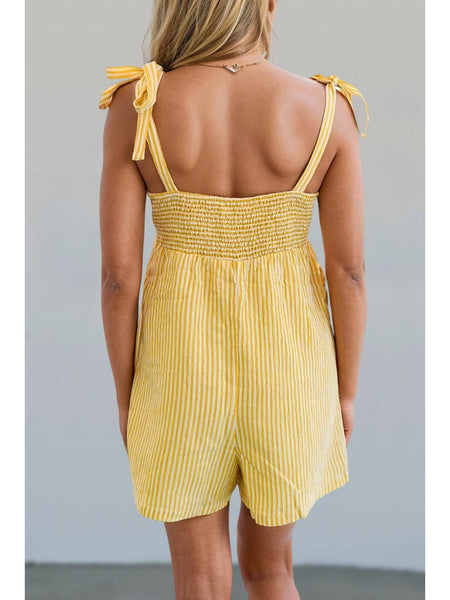 model wearing sleeveless romper in monochromatic yellow vertical pinstripe style pattern with square neckline, tie style wide straps, and relaxed fit high waisted shorts with patch pockets. Shown from the back to display elasticized smocking at back