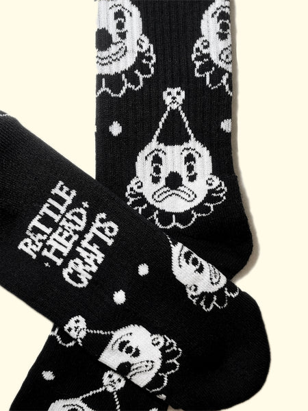 black cotton crew socks with white cuffs and an all over pattern of a white and black clown with either a smiling or frowning face. Shown flat with branding of sock on bottom of foot shown