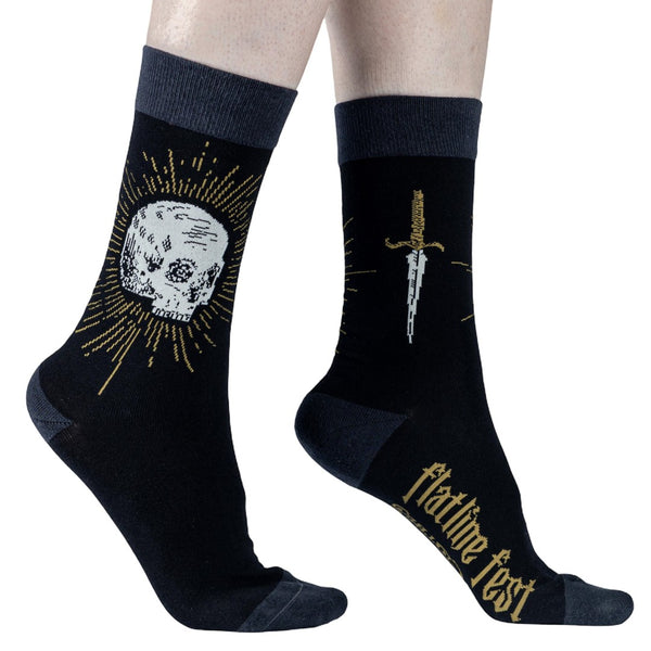 A pair of black cotton crew socks with dark gray cuffs, heels, and toes and design of white skull surrounded by golden yellow sunburst on one ankle and golden handled dagger on the other. Shown worn by model from side