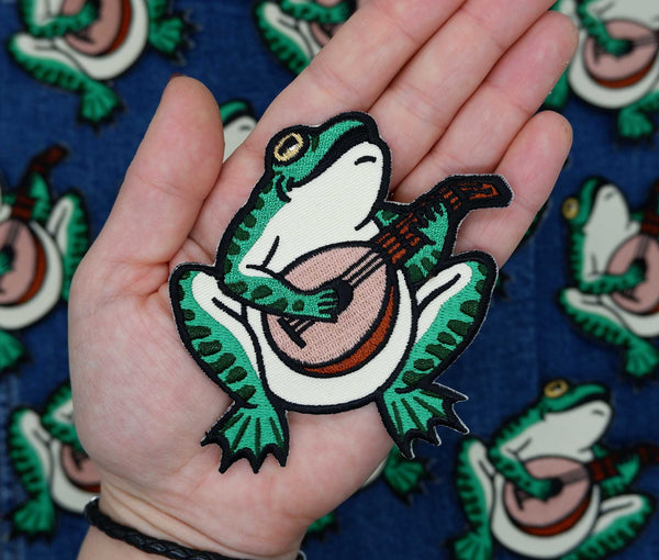 Embroidered patch of a green bullfrog playing a lute and singing. Show held up close