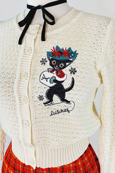 warm white cotton pointelle cardigan with large embroidery on left chest of black cat wearing ice skates, green hat with poinsettias, and white muff alongside trio of snowflakes and "Mischief" written in script below. Shown on dress form from front in close up