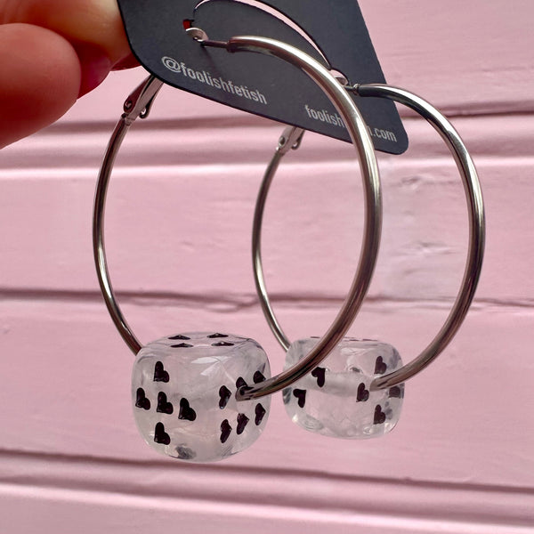 Silver hoop earrings with clear and white marbled dice-shaped charms with black heart-shaped pips on a pink background. Shown from the side