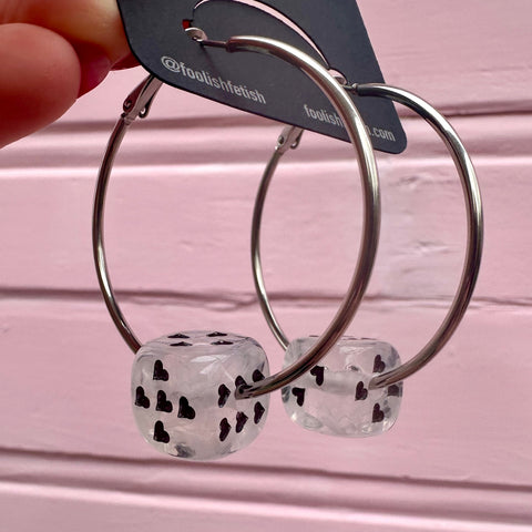 Silver hoop earrings with clear and white marbled dice-shaped charms with black heart-shaped pips on a pink background. Shown from the side