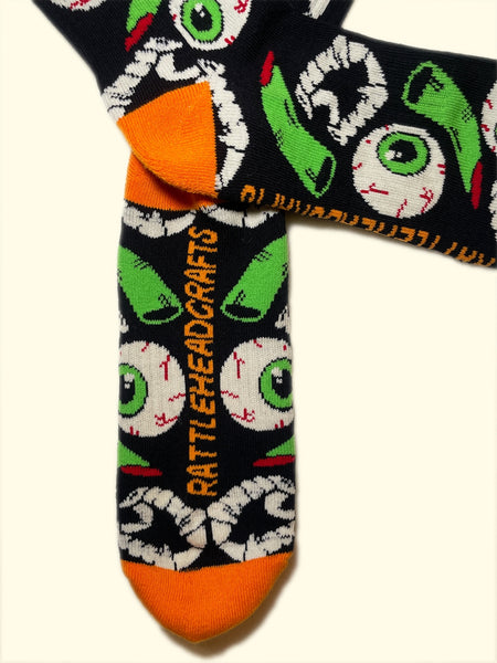 black cotton crew socks with orange cuffs, heels, and toes with an all-over pattern of plastic vampire teeth, witch fingers, and green bloodshot eyeballs. Shown flat with branded bottom of sock shown