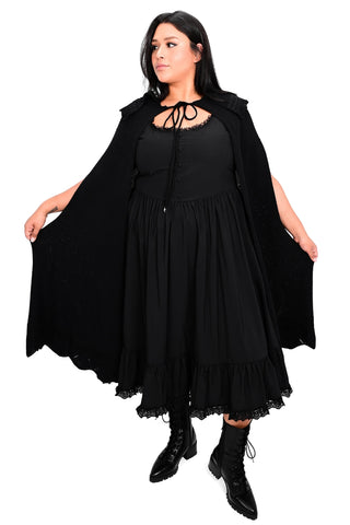 Model wearing black waffle sweater knit cape with scalloped collar and bottom hem with lace detail at collar and bottom hem and black drawstring with silver crescent moon charms. Shown from front