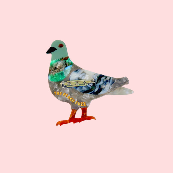 marbled acetate brooch of a pigeon with green head, grey body, and black & white wings with orange legs and feet. Shown from back on pink background